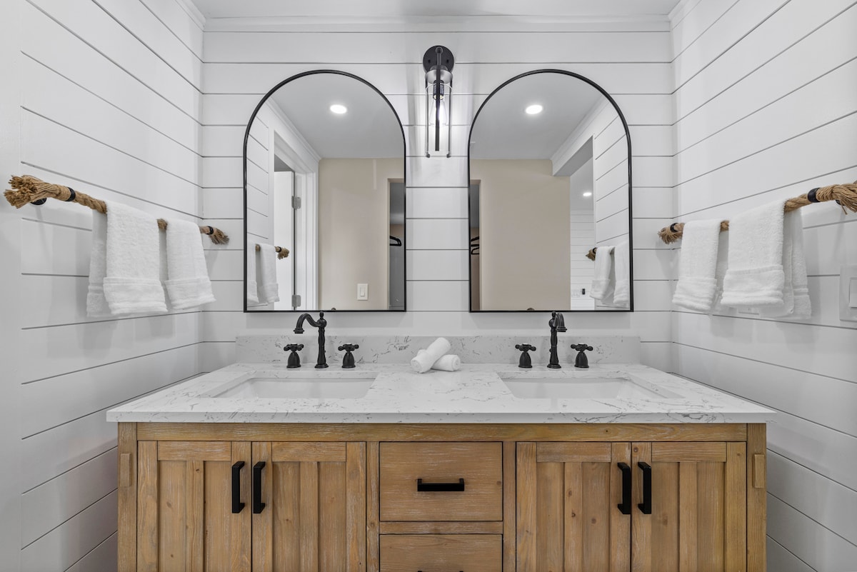 A spacious bathroom features dual sinks with elegant black faucets mounted on a marble countertop. Two large, round mirrors are framed in black, reflecting the light from overhead fixtures. Fresh white towels are neatly hung on wooden rods beside each sink.