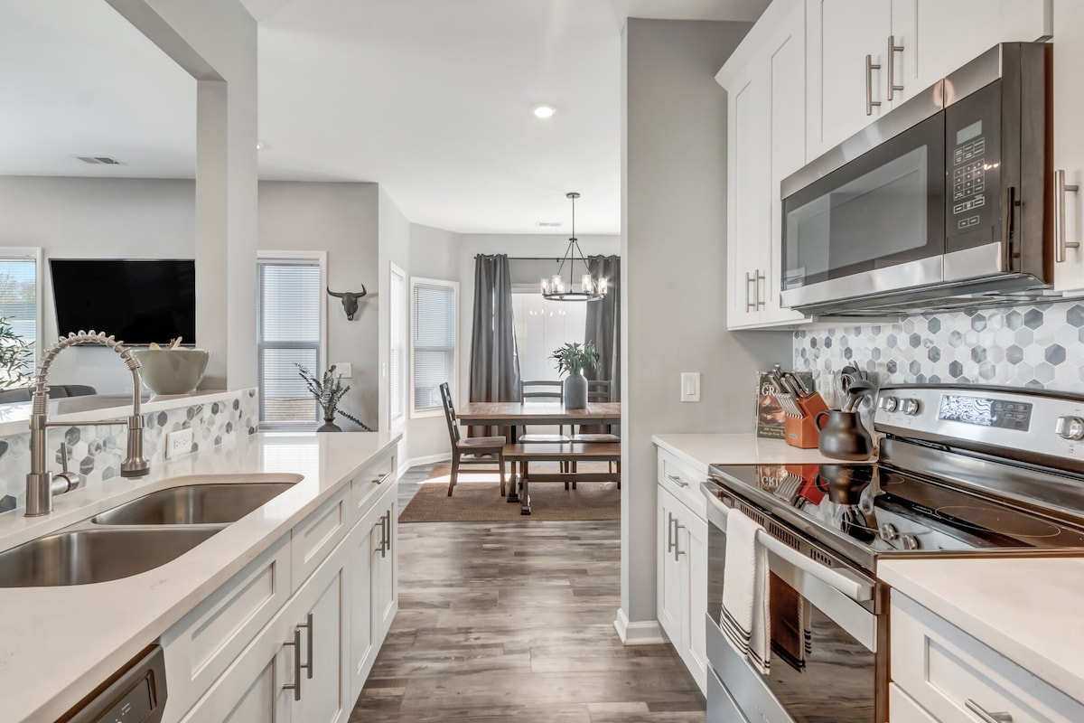 A modern kitchen is highlighted by sleek white cabinetry and stainless steel appliances. A contemporary dining area is visible in the background, featuring a dining table surrounded by chairs. Natural light fills the space, enhancing the open layout.