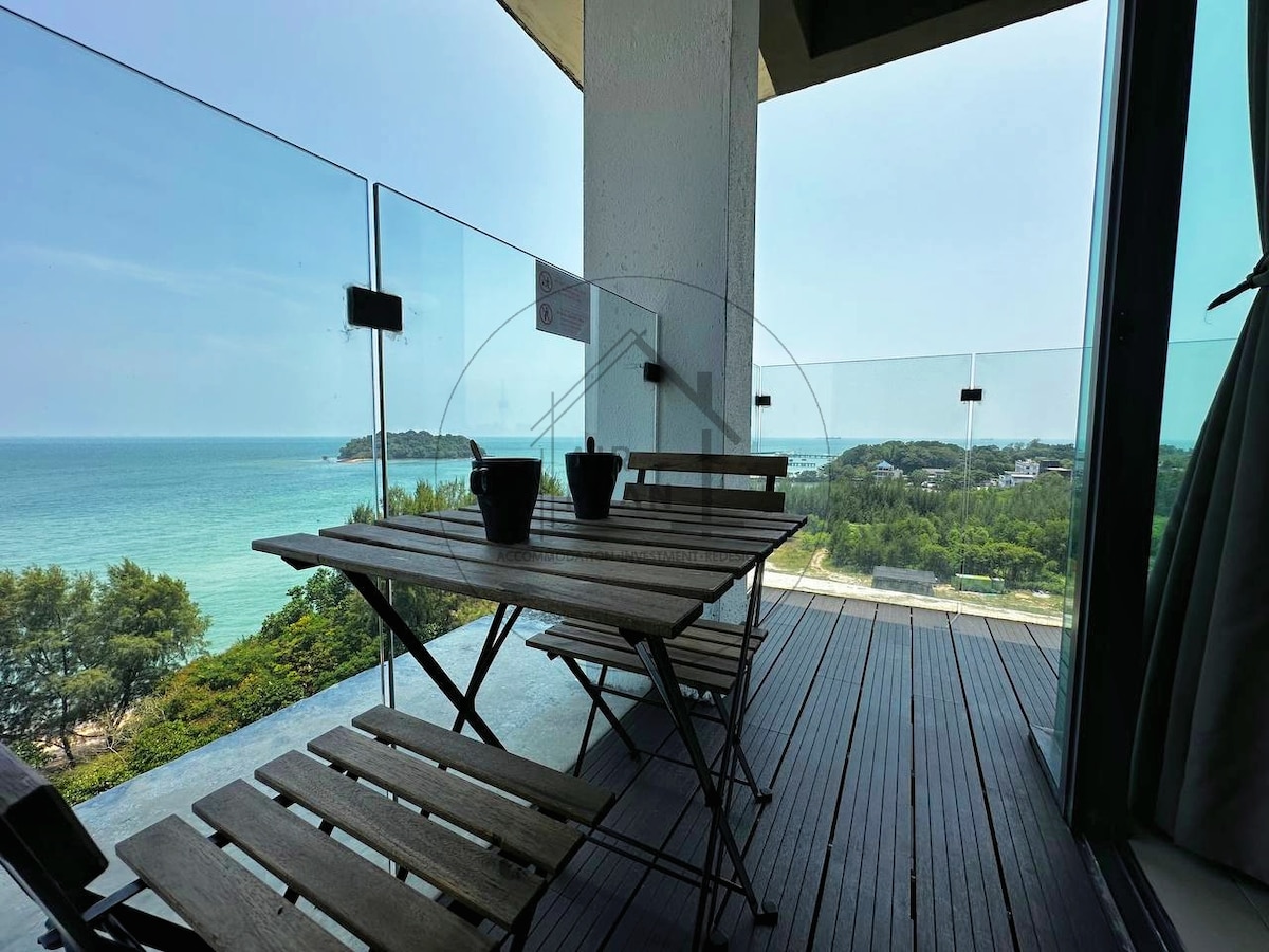 A balcony area features a wooden table and chairs, providing a relaxed seating space. Glass railings offer unobstructed views of the sea and coastal landscape, contributing to an inviting outdoor setting.
