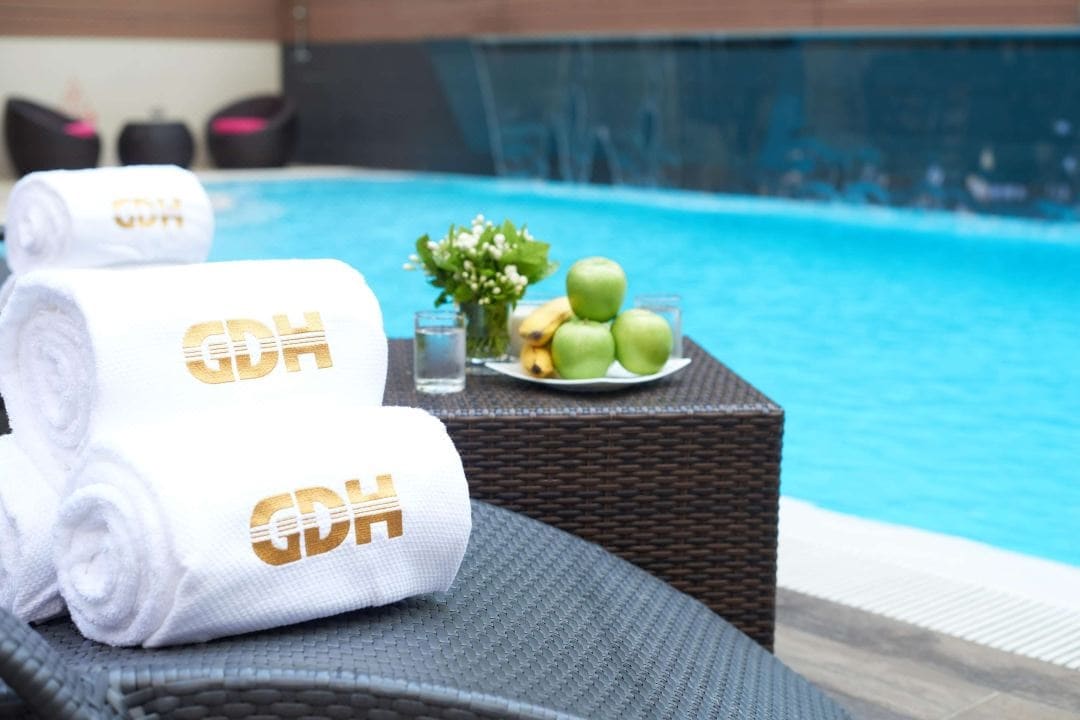 A neatly arranged stack of white towels, embossed with a logo, is placed on a woven lounge chair. A small plate holding green apples and bananas sits nearby, with a backdrop of a clear swimming pool surrounded by a contemporary deck.