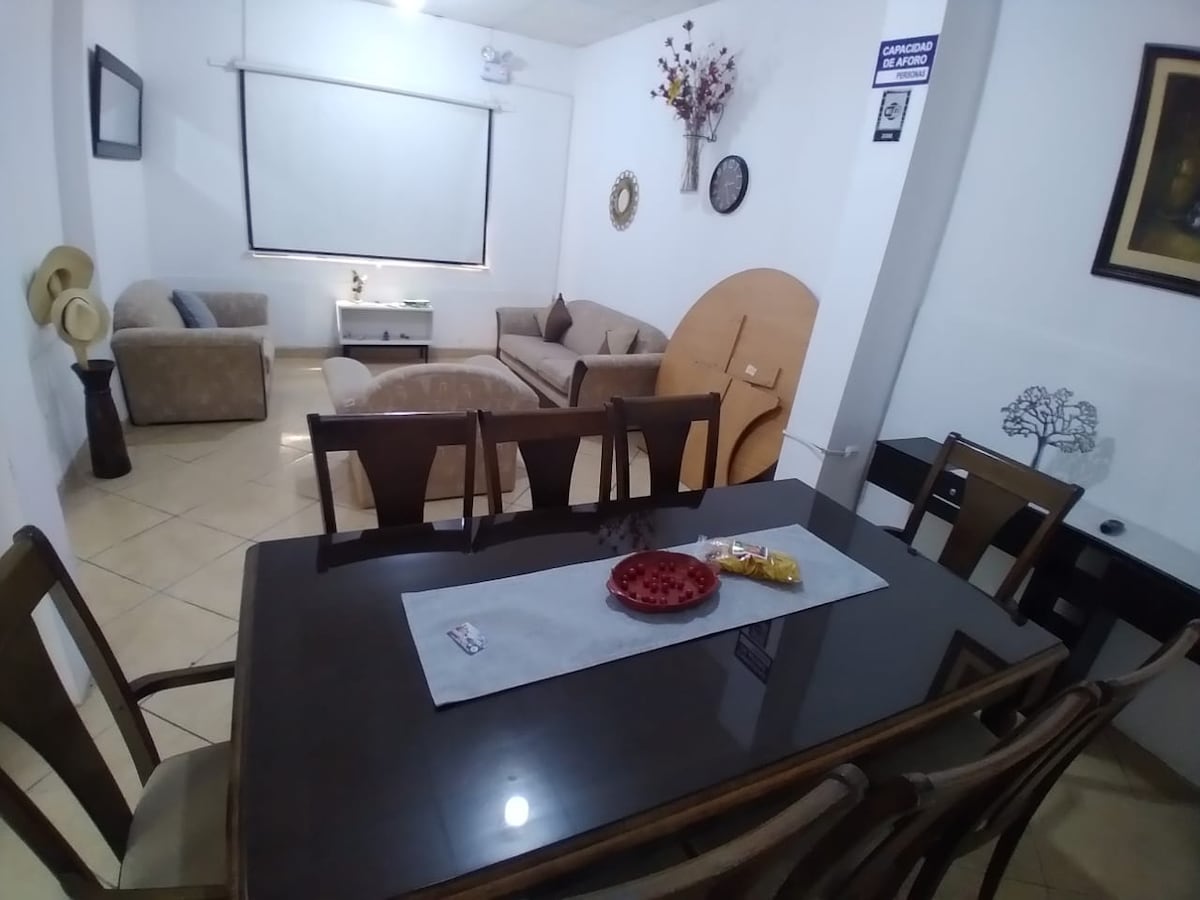 A dining area features a dark wooden table set for six, complemented by matching chairs. In the background, a cozy living area is visible, furnished with soft cushioned sofas. Decorative elements, including a wall mirror and artificial flowers, enhance the overall ambiance.