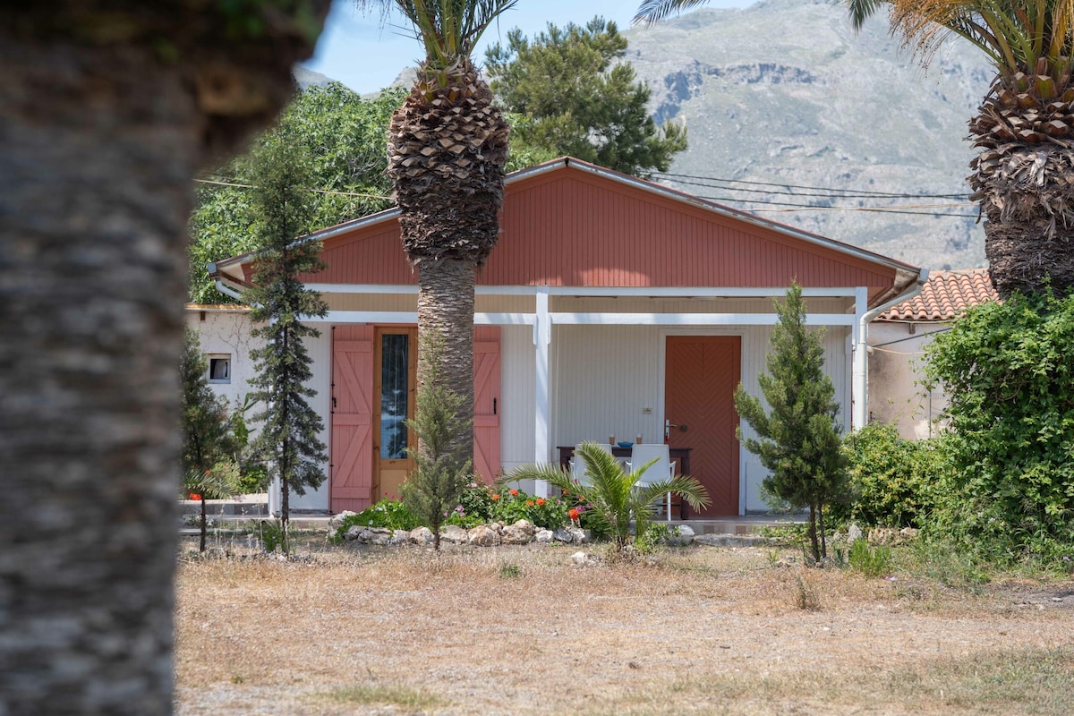 A charming beach house exterior is framed by tall palm trees and lush greenery. A covered porch features coral-colored doors, welcoming visitors. The natural landscape includes dry grass and surrounding vegetation, with mountains visible in the distance.