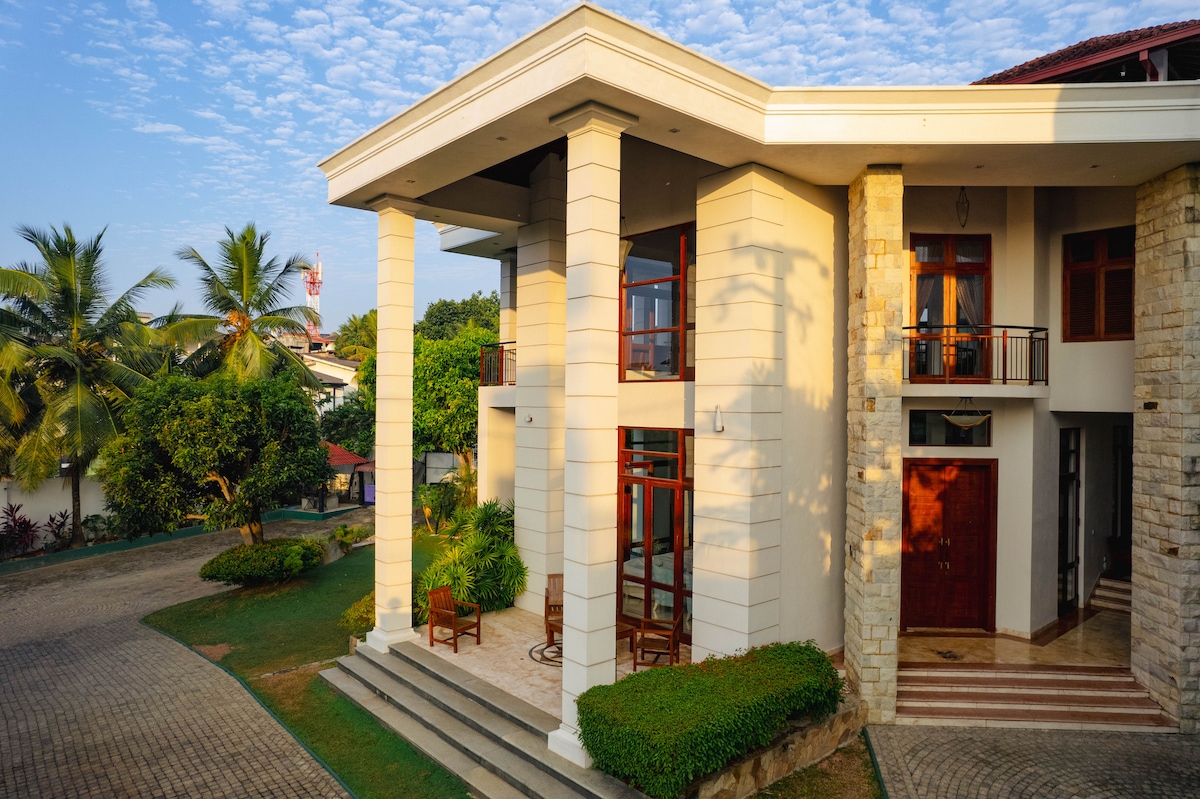An elegant two-story villa is set amidst green landscapes. The façade features tall columns and large glass doors that open to a spacious entrance. Surrounding the villa, well-maintained gardens with palm trees and shrubs provide a serene environment.