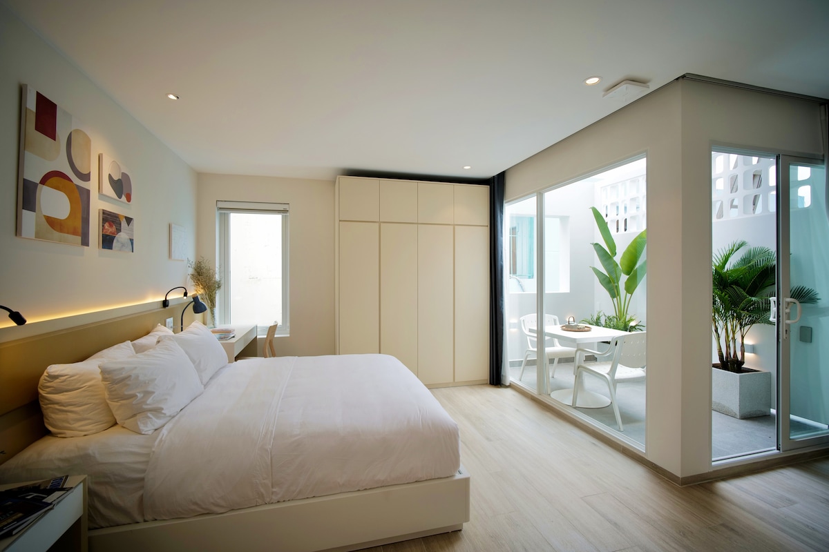 A modern studio apartment features a spacious bed, minimalistic furniture, and a large window leading to a private balcony with greenery. Light wooden flooring complements the soft colors of the walls, and natural light enhances the inviting atmosphere. Artwork is displayed above the nightstand.