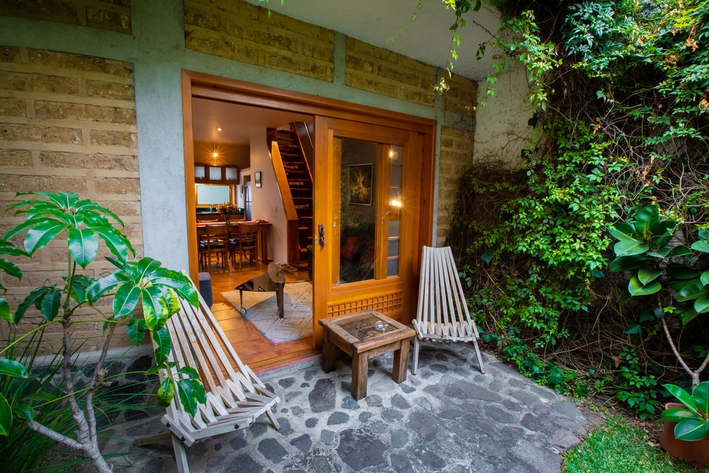 A small outdoor terrace features two wooden lounge chairs positioned beside a rustic table. Lush greenery surrounds the area, along with stone flooring that provides a natural aesthetic. The entrance to the interior space is framed by large glass doors, inviting light into the home.