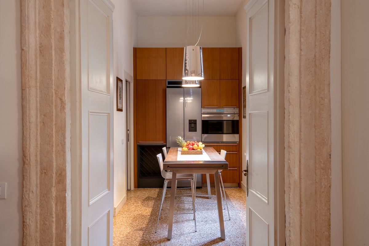 A modern kitchen is shown, featuring wooden cabinetry and stainless steel appliances. A central dining table is surrounded by four minimalist chairs. Soft lighting highlights the space, while the open doorway creates a flow from the kitchen to the adjoining areas.