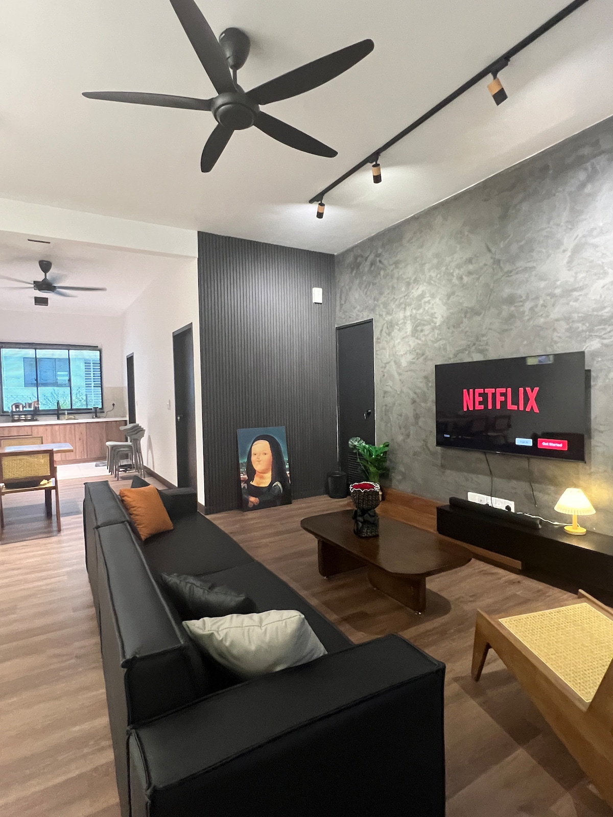 A modern living area is furnished with a dark sectional sofa, complemented by a small coffee table. A wall-mounted TV displays the Netflix logo, while light fixtures illuminate the space. A dining area is visible in the background, featuring a table and chairs by a large window.