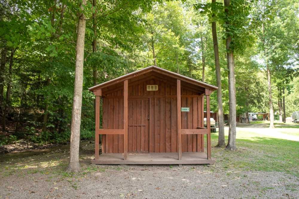 A rustic cabin is nestled among trees, featuring a wooden facade with a simple overhang. The entrance is framed by wooden support beams, and a gravel path leads to the front, surrounded by a natural landscape.