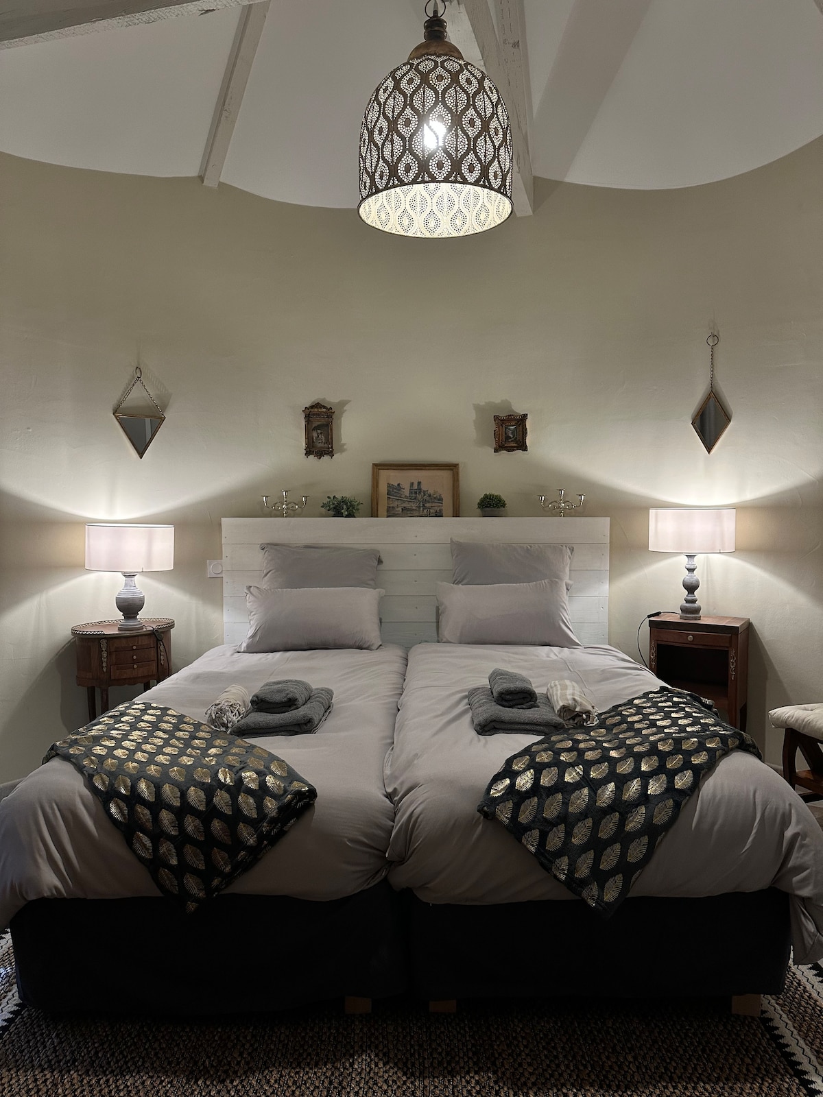 A cozy bedroom is showcased, featuring two spacious beds with muted gray linens and decorative throws. Soft lighting is provided by two bedside lamps, and wall art adds subtle elegance. A woven rug lies beneath, enhancing the ambiance of the room.