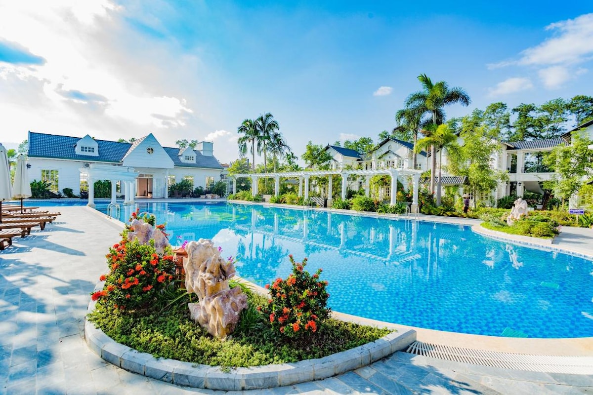 A large swimming pool is surrounded by lush gardens, featuring vibrant flowers and palm trees. Sun loungers are arranged poolside, inviting relaxation. In the background, a series of charming villa-style buildings are visible, enhancing the tranquil atmosphere.