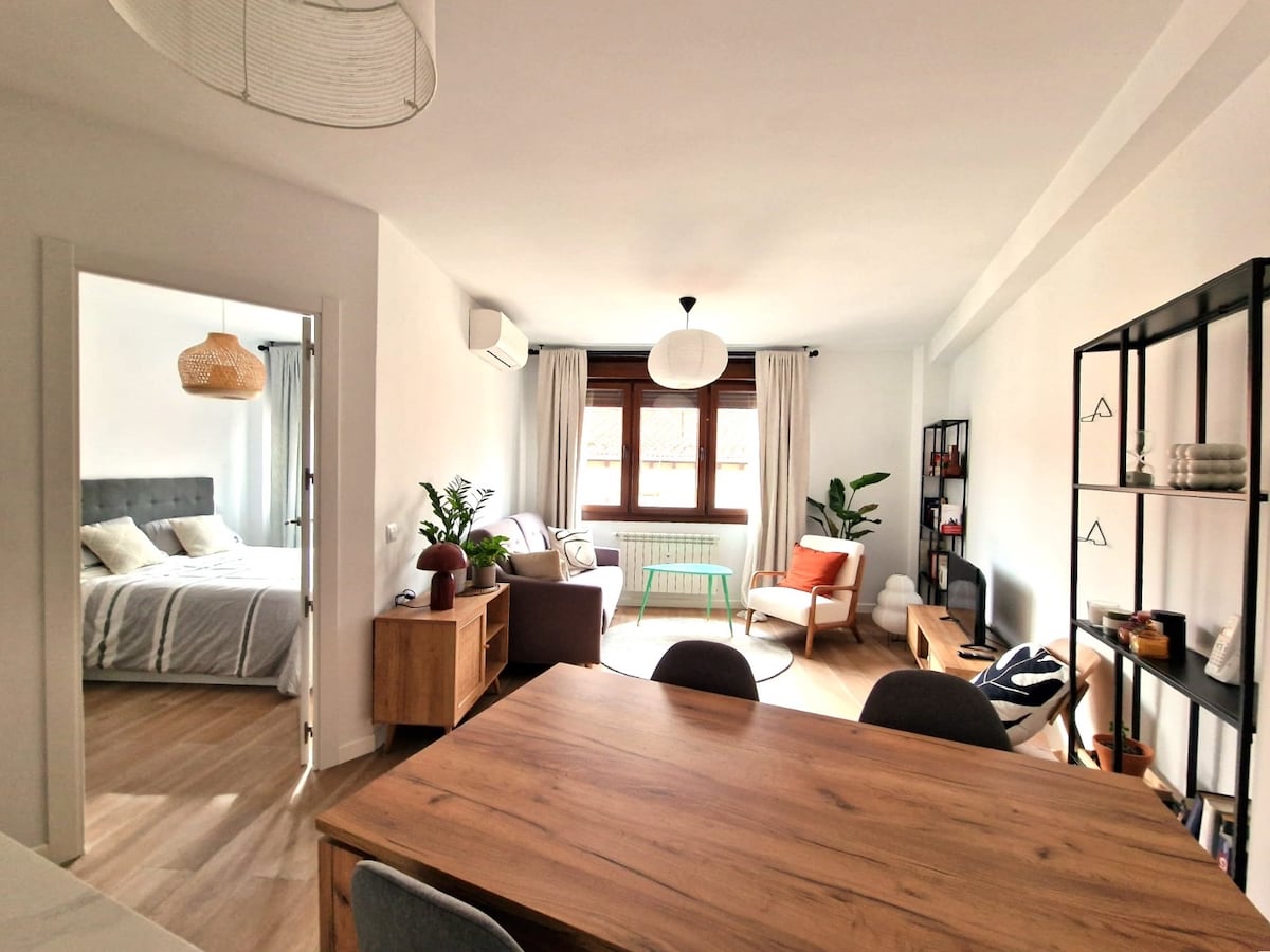 A bright, open-plan living area showcases a wooden dining table accompanied by grey chairs. A comfortable armchair sits near a large window, with plants adding a touch of greenery. The adjacent bedroom is visible through an open door, featuring a soft bed and natural light.