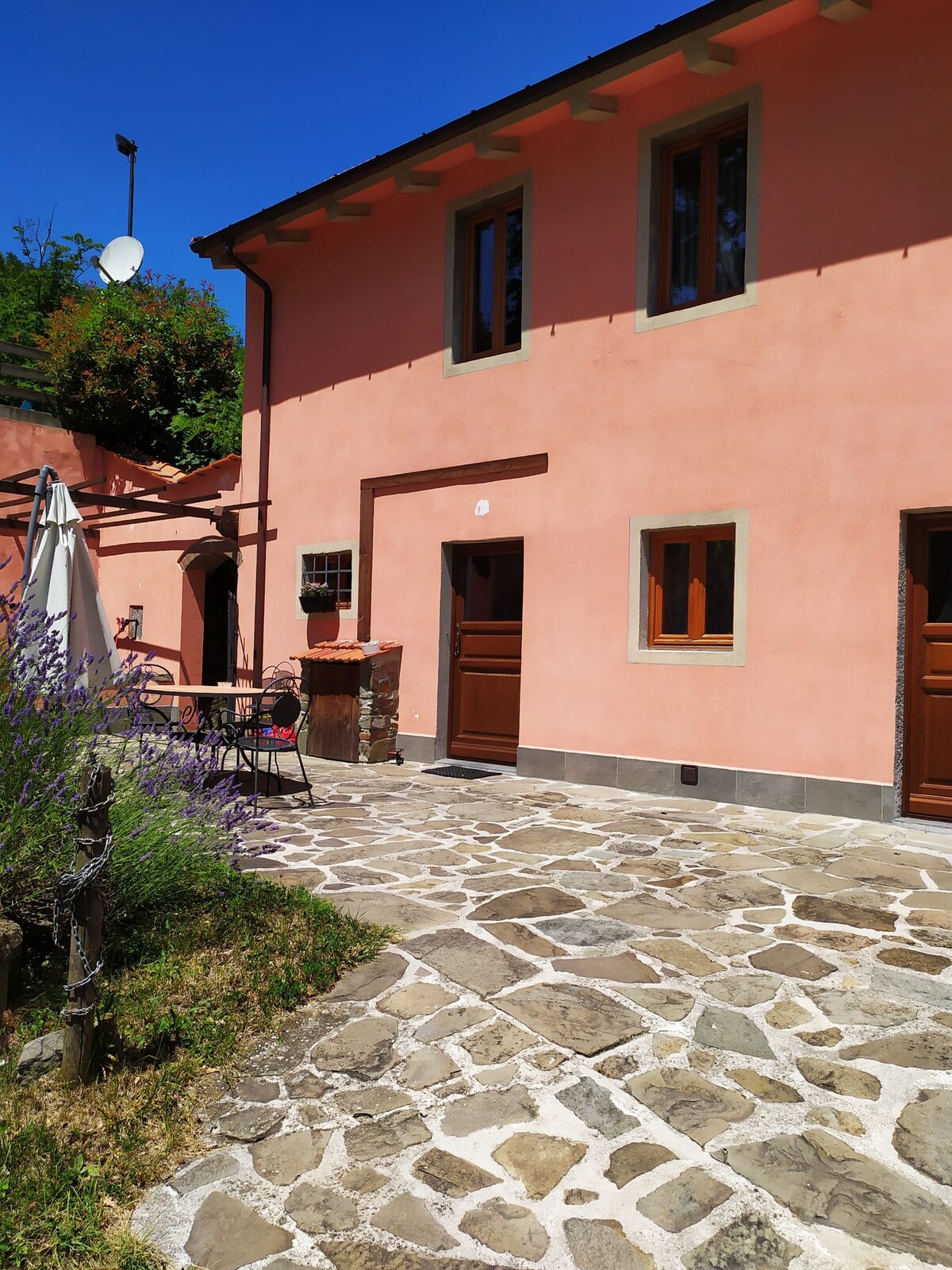 The exterior of a charming, pink-hued villa is presented, featuring stone pathways and lush greenery. Outdoor seating is provided with a small table and chairs, complemented by a parasol. Windows are framed in a neutral tone, with an inviting entrance visible.