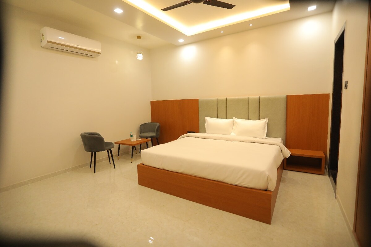 A modern room features a spacious bed with crisp linens, flanked by soft gray chairs and a small table. Warm lighting accentuates the minimalist design, and an air conditioning unit is mounted on the wall.