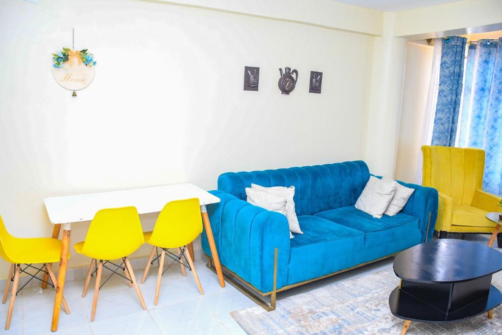 A living area features a blue sofa accompanied by two yellow chairs and a white dining table. Decorative items are placed on the wall, and large windows with sheer curtains provide natural light.