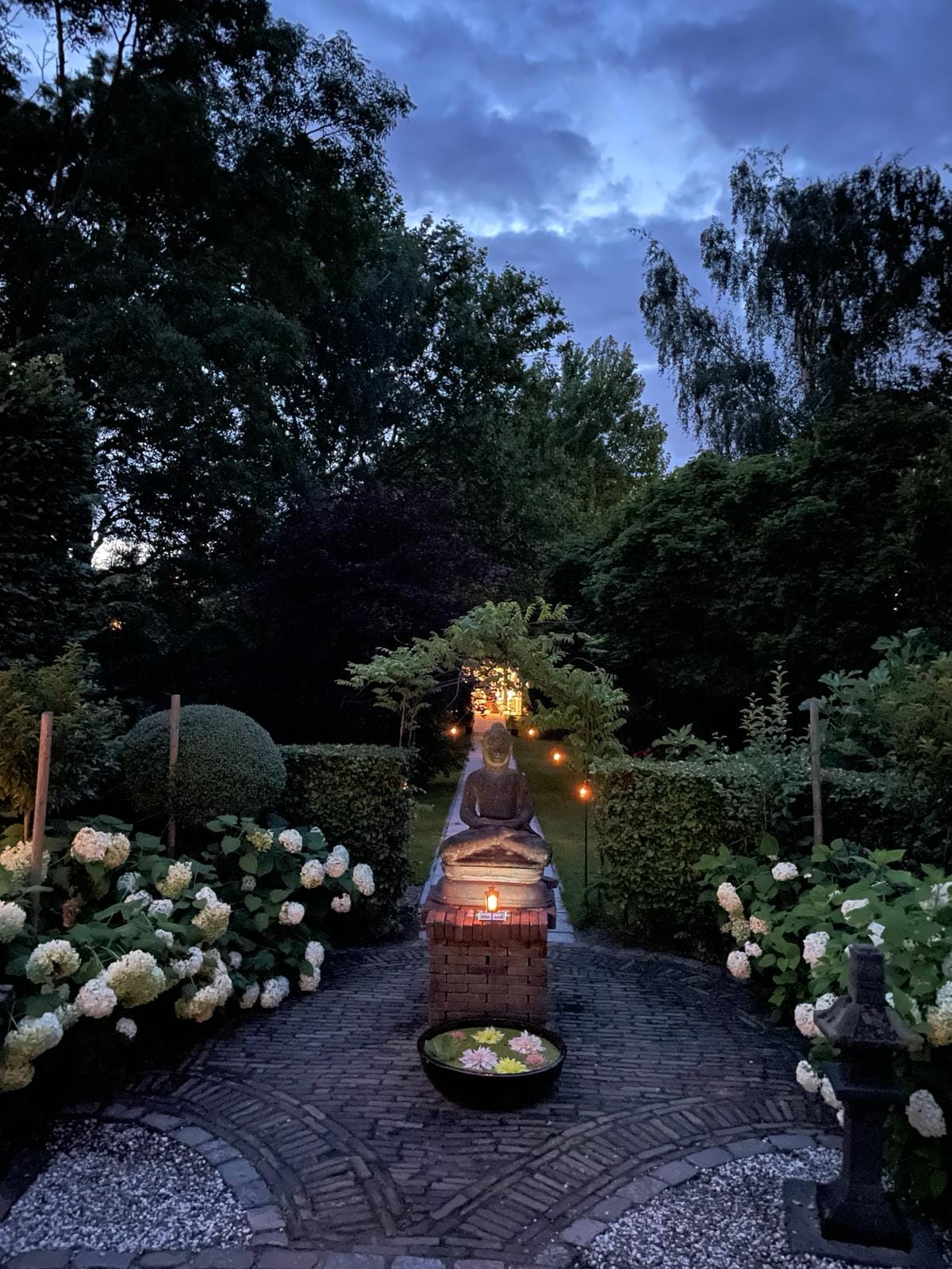A tranquil garden scene is illuminated by soft lighting, showcasing a stone statue at the center. Lush greenery surrounds winding paths, lined with blooming hydrangeas. The evening sky casts a gentle blue hue, enhancing the serene atmosphere.