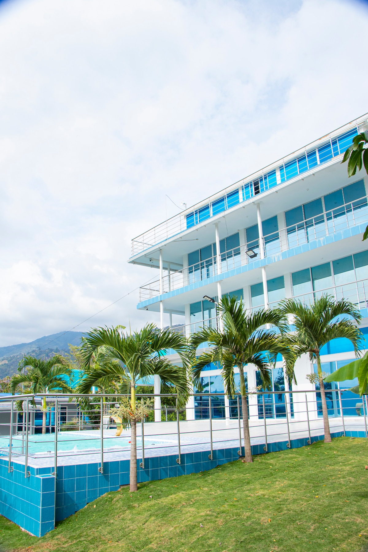 A modern building features expansive glass balconies and is surrounded by lush palm trees. A tranquil swimming pool, set against a backdrop of gentle hills, provides a refreshing escape. Spacious outdoor areas are ideally designed for relaxation in a serene environment.