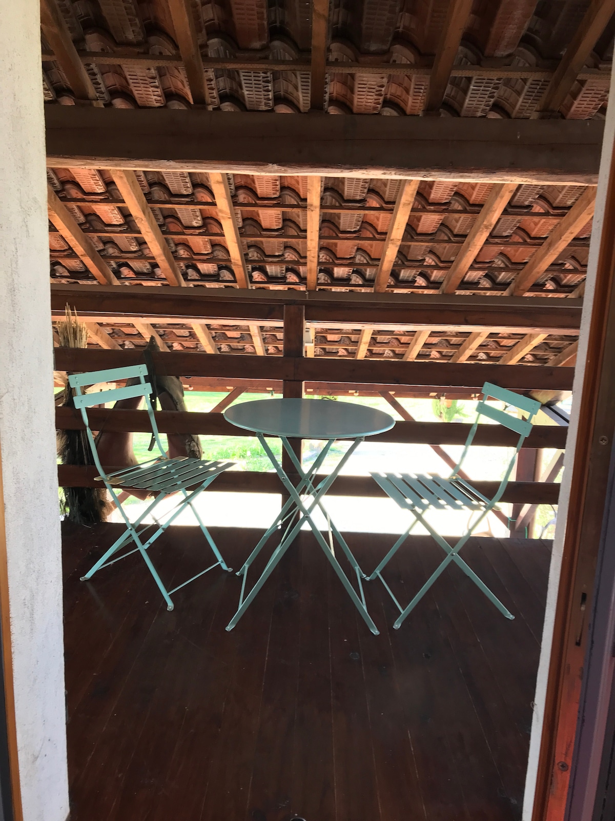 A balcony space is presented with a small round table and two folding chairs. The wooden flooring is visible beneath, and the ceiling features exposed beams, creating an open feel. A view of greenery can be seen through the balcony railing.