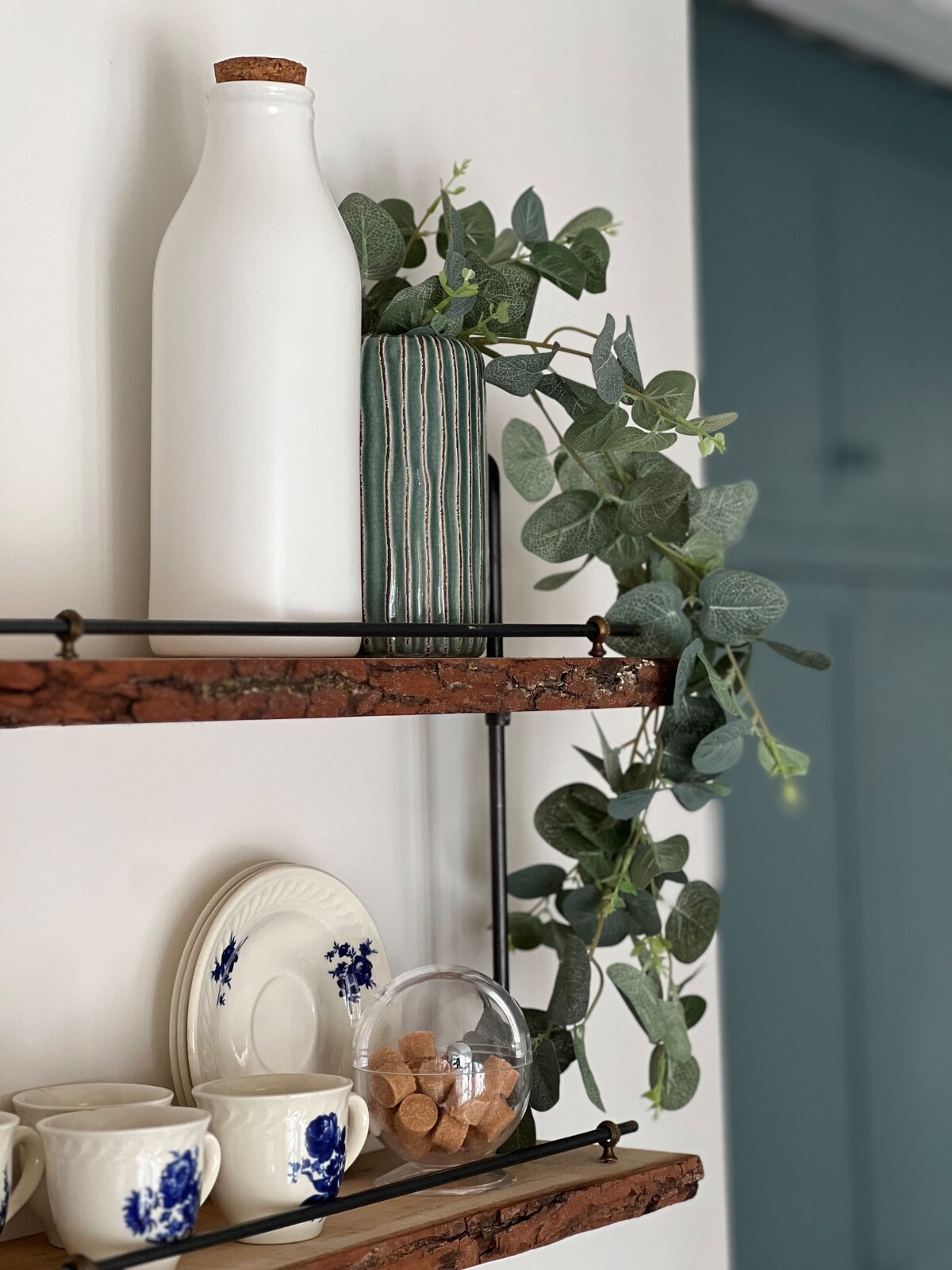 A shelf displays a white ceramic bottle and a striped green vase, accompanied by artificial greenery. Below, delicate blue and white cups rest alongside a glass jar filled with sugar cubes, creating a charming, organized arrangement.