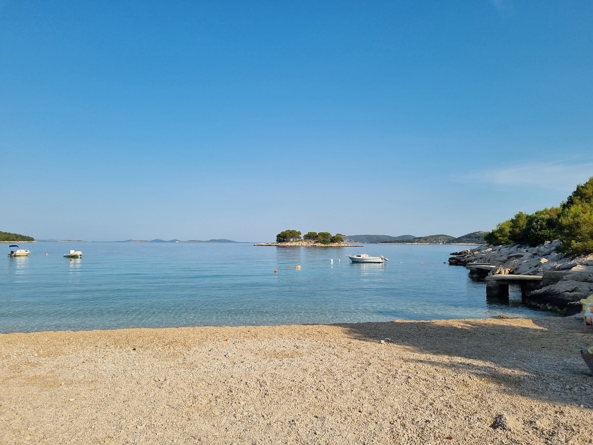 A serene beach scene is presented, featuring calm waters reflecting a clear blue sky. Several small boats are anchored nearby, and a rocky shoreline is visible. Lush green trees can be seen in the distance, offering a sense of tranquility to the coastal setting.