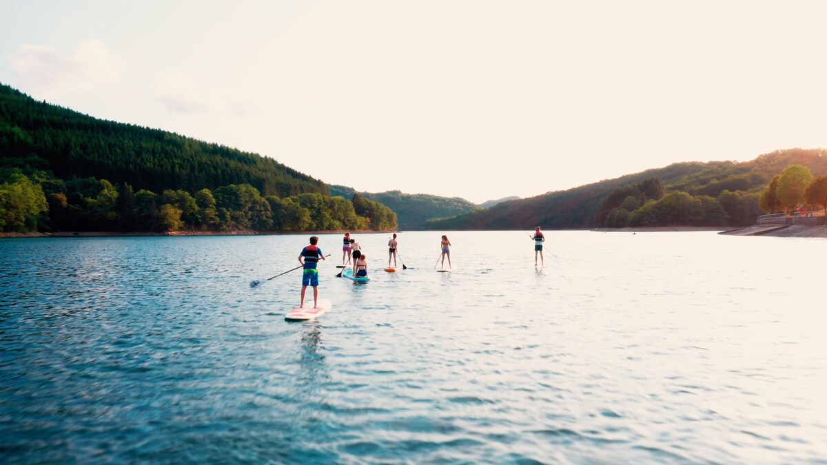 A group of individuals paddle boarding on a serene lake, surrounded by lush green hills. The water reflects the soft light of the setting sun, creating a peaceful atmosphere. The shoreline is visible in the background, enhancing the natural beauty of the location.
