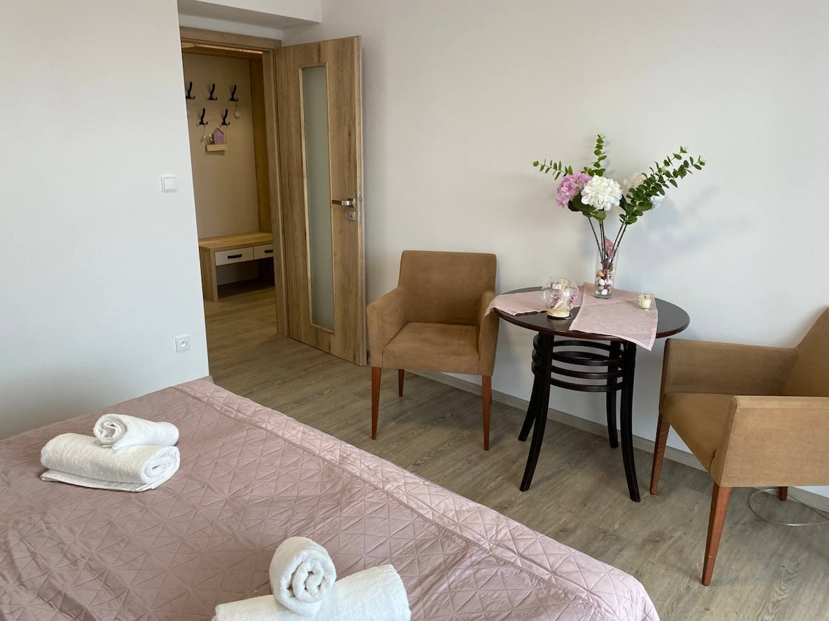 A cozy room displays a spacious bed adorned with neatly rolled towels. Two brown chairs are positioned around a small, dark wooden table, which is set with a pink cloth and a floral arrangement. A door leads to another area, complementing the light floor and neutral wall colors.