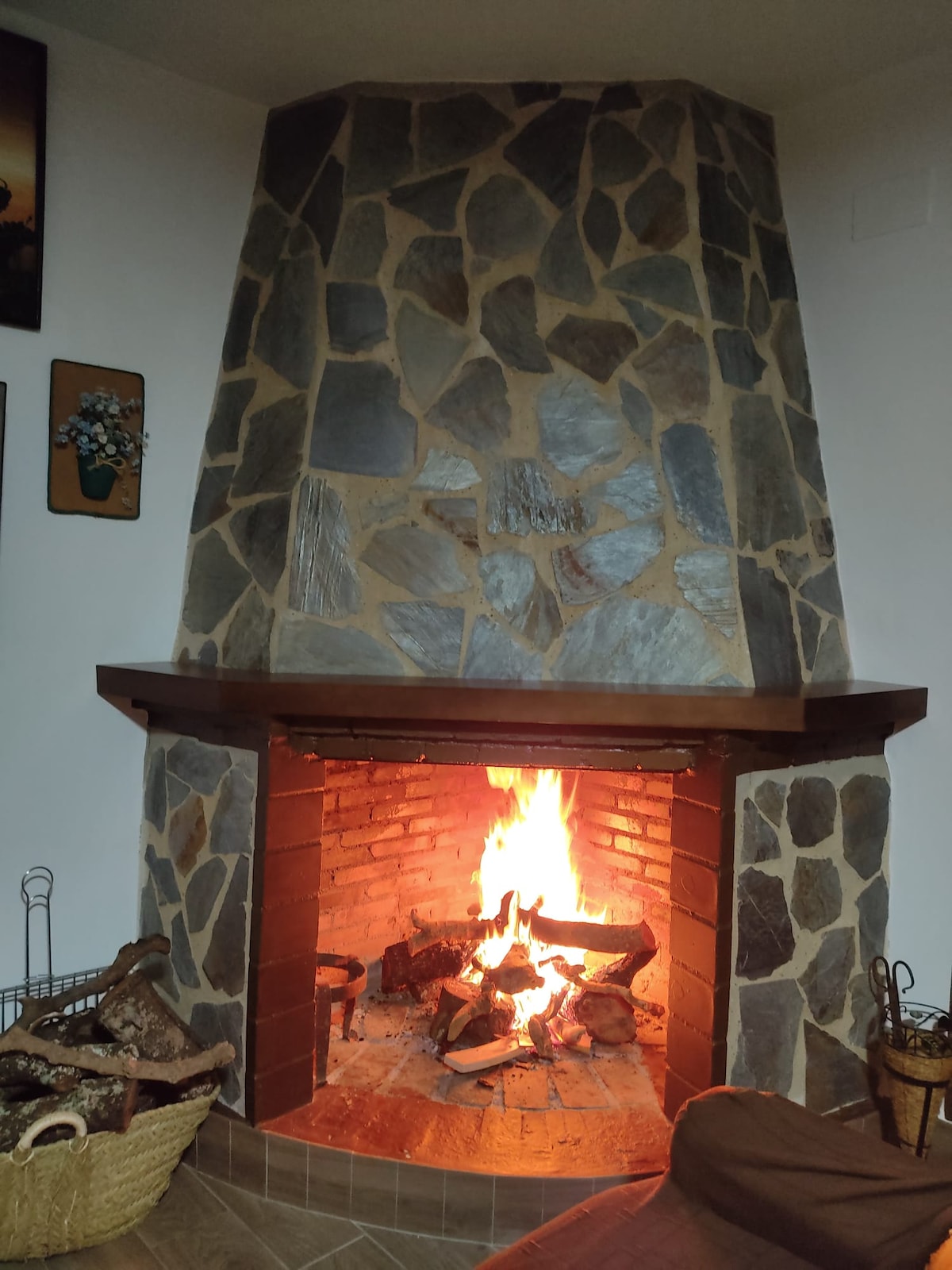 A rustic stone fireplace features a roaring fire, surrounded by neatly stacked logs. The hearth is enhanced by a wooden mantel, while the warm glow of the flames contrasts with the cool tones of the stone, creating a cozy and inviting ambiance.