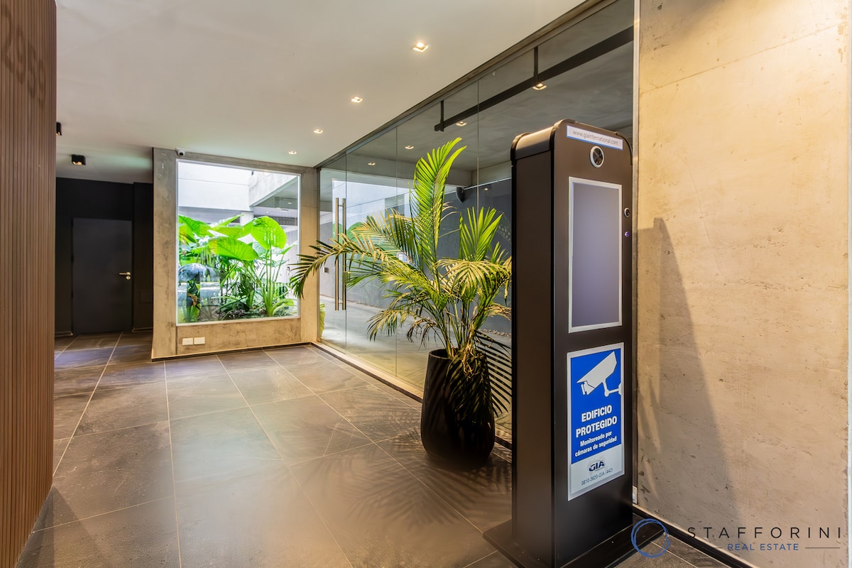 A modern lobby area features sleek dark tile flooring and large glass panels that allow natural light to fill the space. Lush green plants add a touch of nature, complementing the contemporary design. A security totem is visible, enhancing the sense of safety.