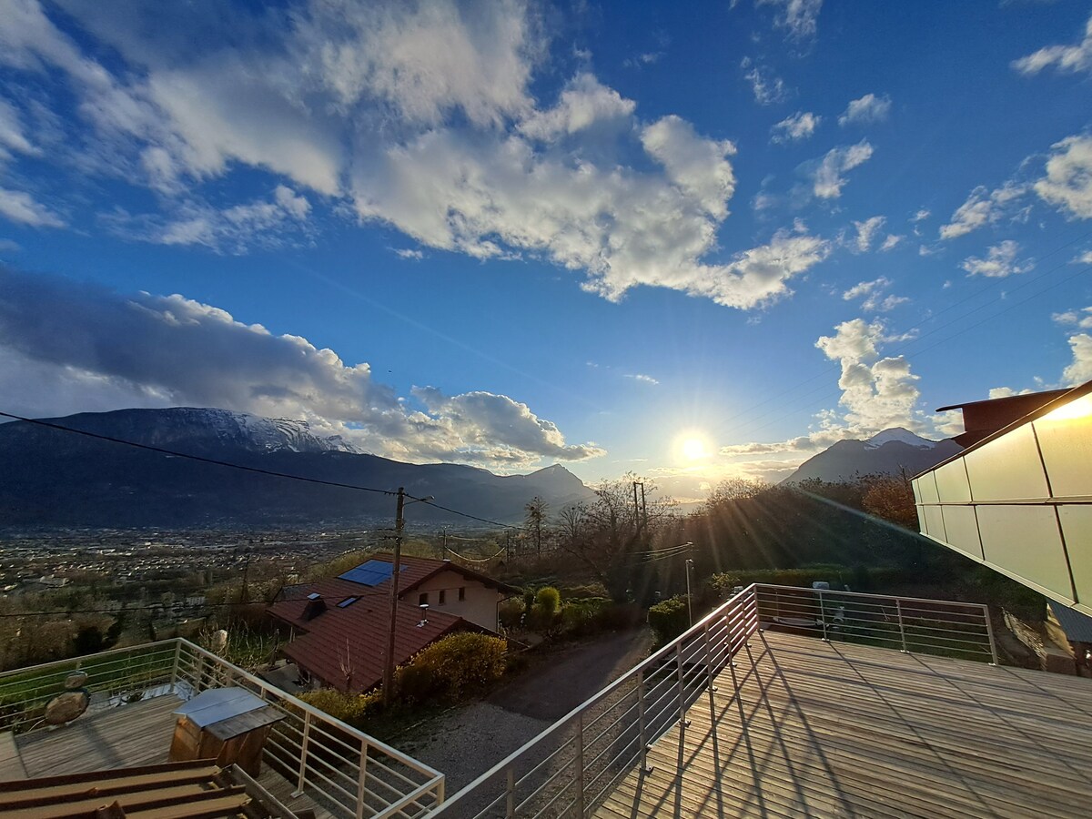 A scenic view of mountains and valleys is captured at sunset, with the sky featuring an array of clouds in soft colors. A spacious terrace is visible in the foreground, providing an inviting space to enjoy the surroundings.