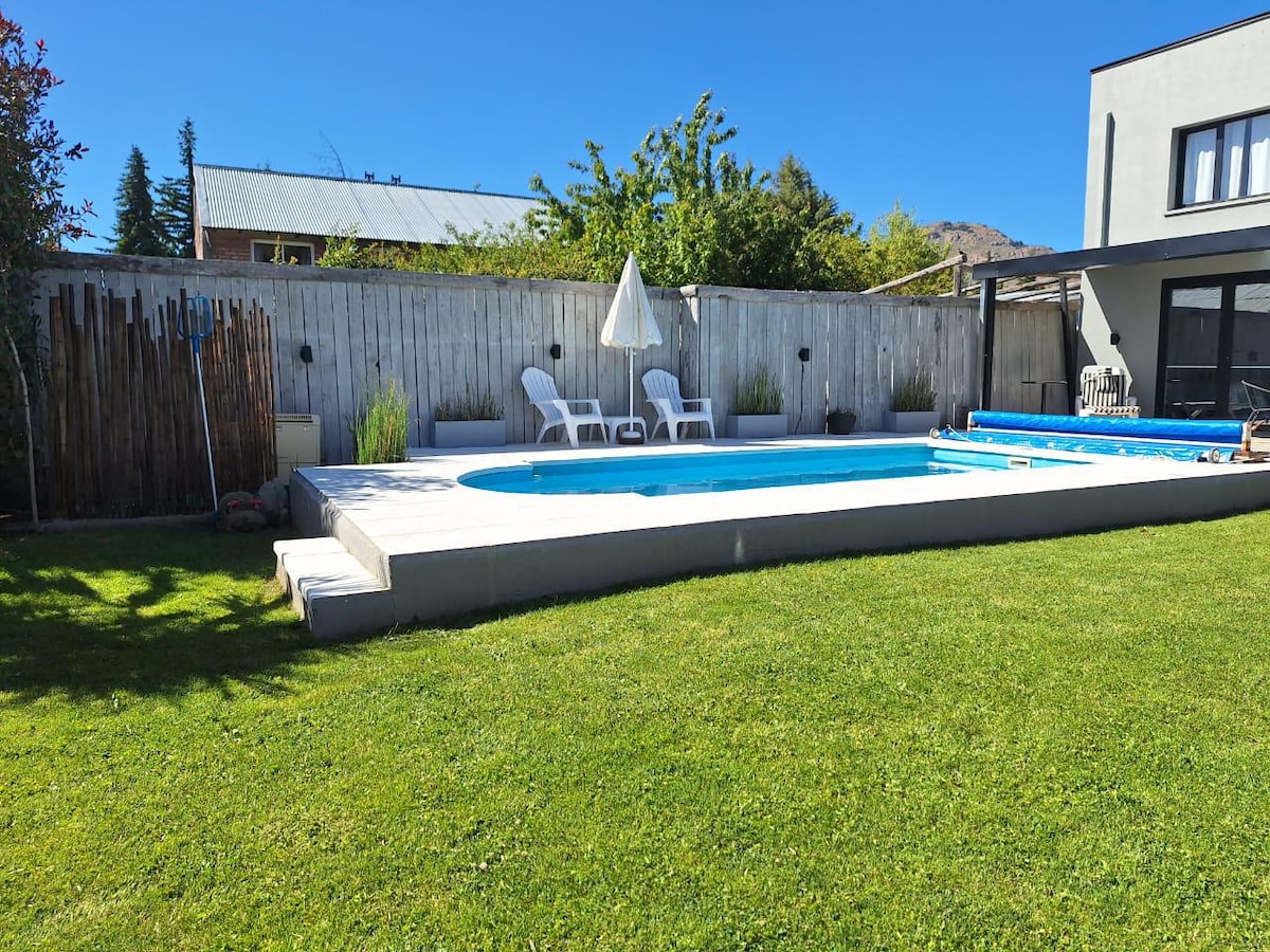 A well-maintained outdoor space features a heated swimming pool surrounded by a grassy area. Two lounge chairs offer seating, and a large umbrella provides shade. The landscaping includes low shrubs and a wooden fence, enhancing the tranquil environment of the patio area.