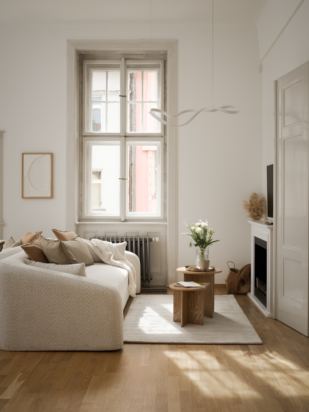 A bright living area features a cozy, textured sofa adorned with soft pillows. Natural light filters through an elegant, large window, enhancing the serene atmosphere. A simple wooden coffee table supports a vase of fresh flowers, while a minimalist fireplace complements the space.