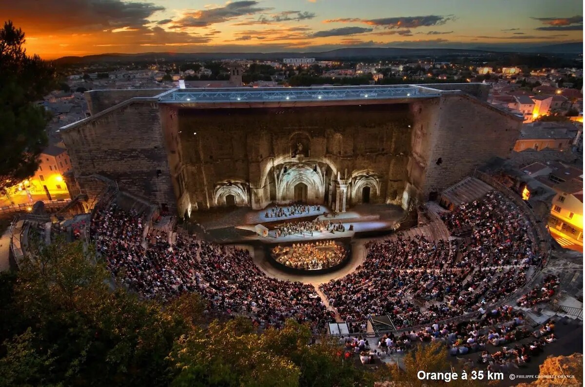 An outdoor amphitheater is seen, filled with a large audience attending a performance. The backdrop features ancient stone walls illuminated by the warm hues of sunset, enhancing the scenic view. A stage occupies the central area, where performers can be seen.