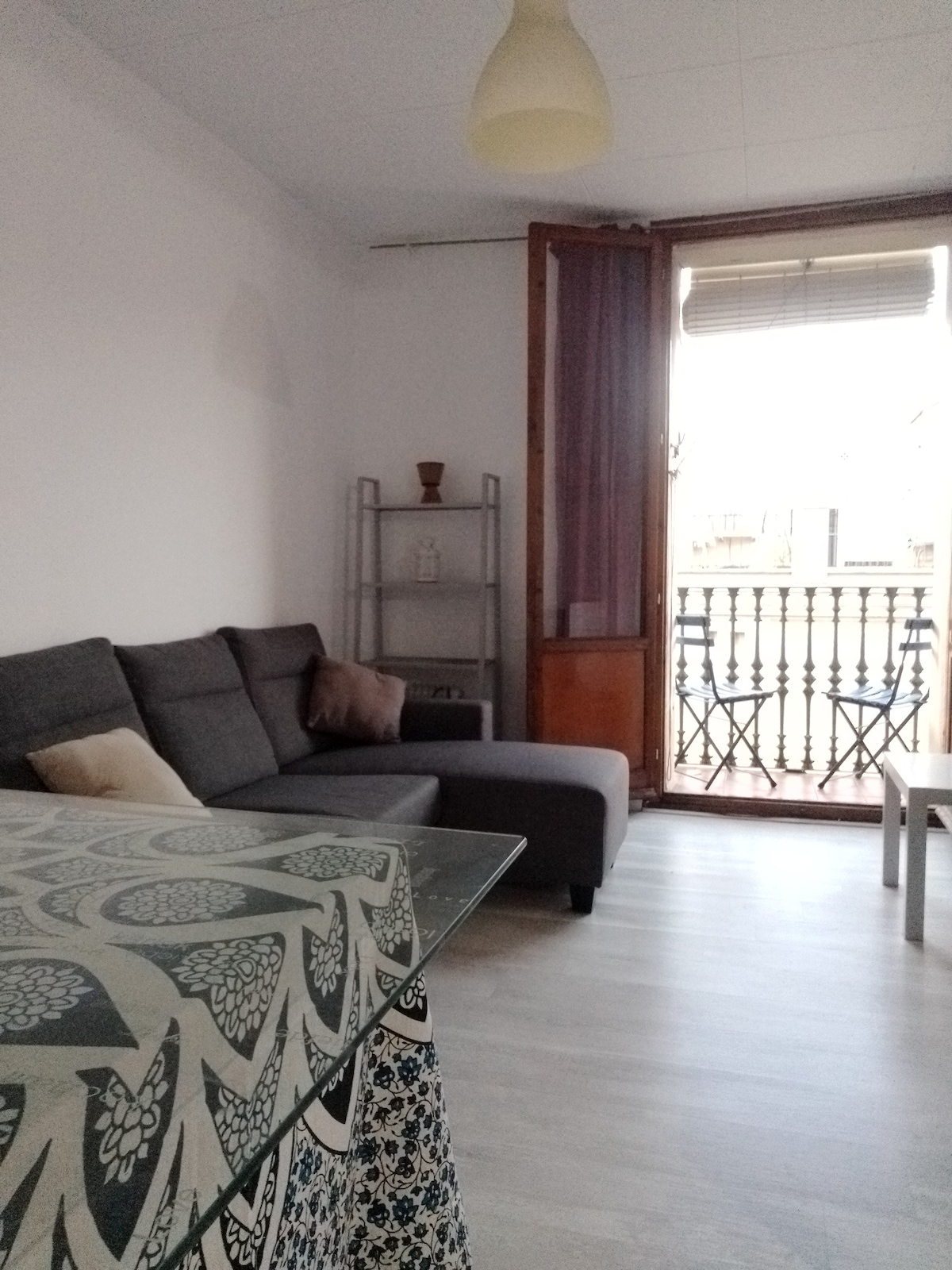 A comfortable living area is displayed, featuring a gray sectional sofa and a glass dining table adorned with a patterned tablecloth. Natural light floods through the large window with wooden shutters, which opens to a small balcony overlooking the street.