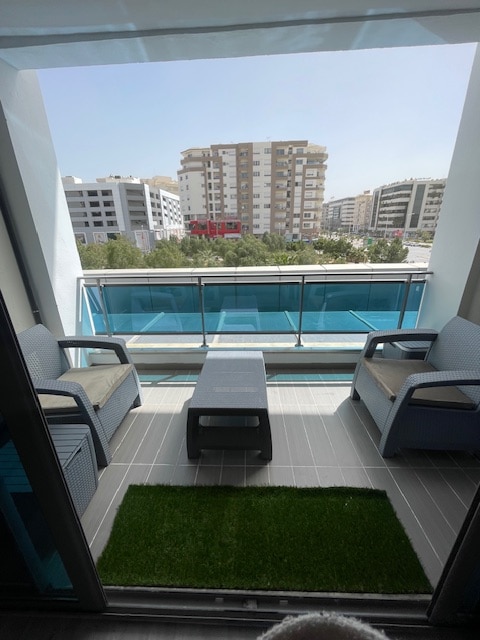 A private balcony is visible, featuring two wicker chairs and a low table. A small patch of artificial grass enhances the space. The surrounding view includes modern buildings and greenery, allowing for an open and bright atmosphere.