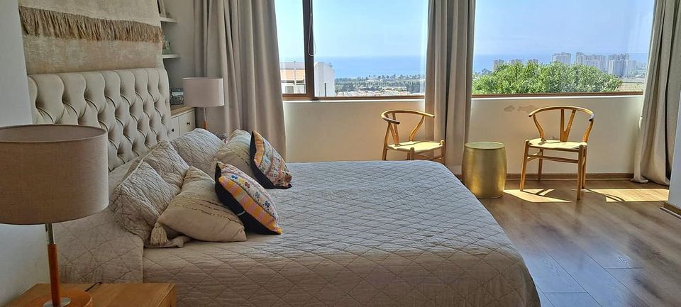 A bright and airy bedroom is presented, featuring a spacious bed dressed in neutral tones and adorned with patterned pillows. Two wooden chairs are positioned near the window, providing a view of the ocean and nearby landscape. Soft natural light filters through light curtains.