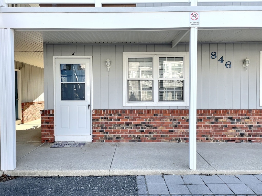 The exterior of the ground-floor condo is visible, featuring a light gray facade with brick accents. A white door marked '2' leads into the unit, flanked by two windows. A concrete pathway offers easy access, sheltered by an overhead awning.