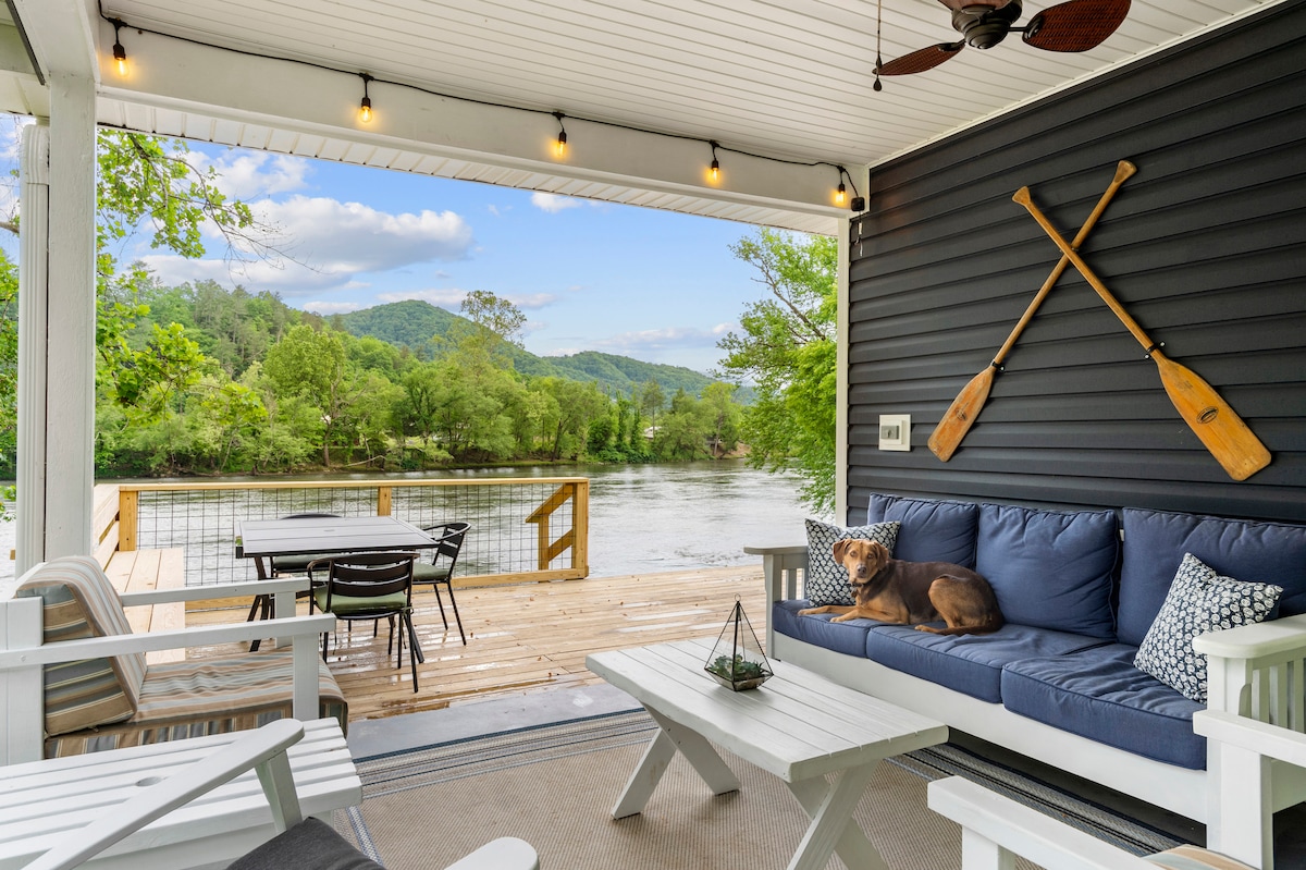 A covered patio area features a comfortable seating arrangement with blue cushions and decorative pillows. A rustic wooden table and chairs can be seen alongside a scenic view of the river and mountains. Two paddles hang on the wall, enhancing the outdoor atmosphere.