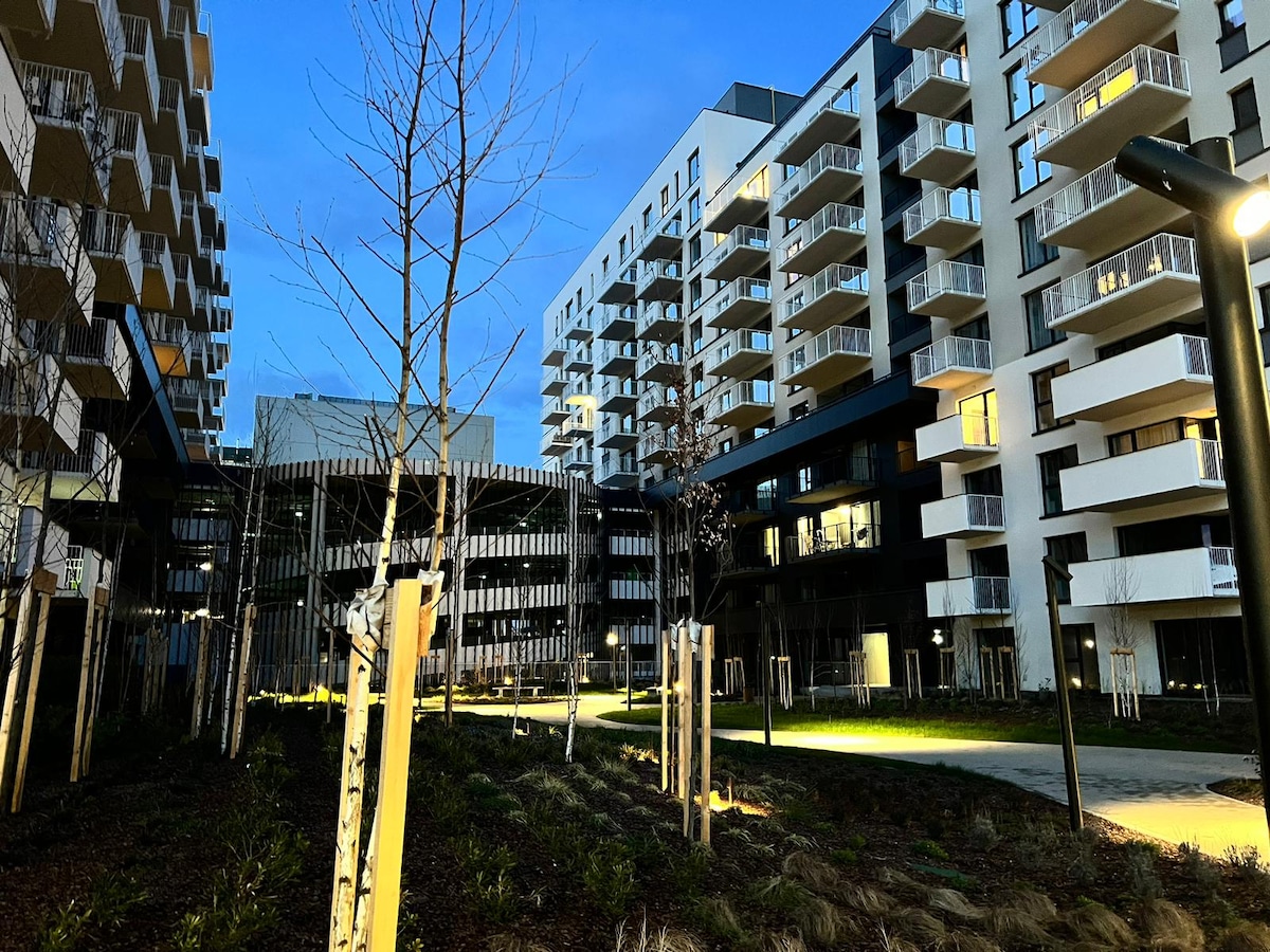 A modern residential complex is captured at dusk, with illuminated balconies and landscaped grounds. Neatly arranged trees and green spaces complement the buildings, creating a serene atmosphere. Soft lighting highlights pathways, enhancing the welcoming environment of the community.