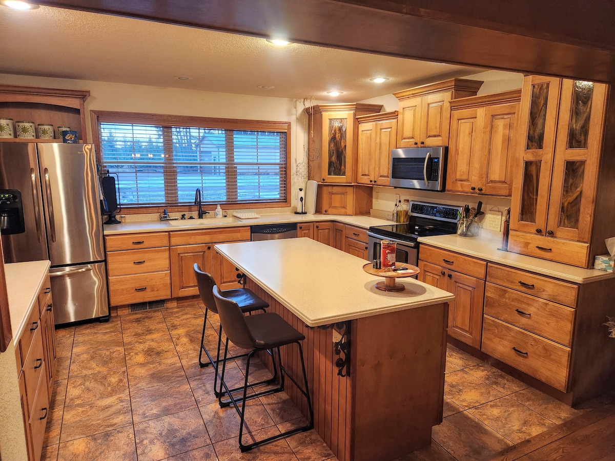 A spacious kitchen is presented with wooden cabinetry and ample counterspace. Stainless steel appliances, including a refrigerator and oven, are visible. A large window allows natural light to fill the area, while modern bar stools are positioned at the kitchen island.