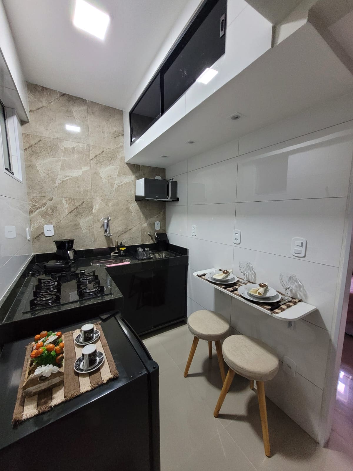The kitchen features modern black cabinetry and a sleek countertop. Essential appliances, including a microwave and stovetop, are readily available. A small dining area with two stools is positioned adjacent to the wall, while a decorative tray with snacks is arranged on the counter.