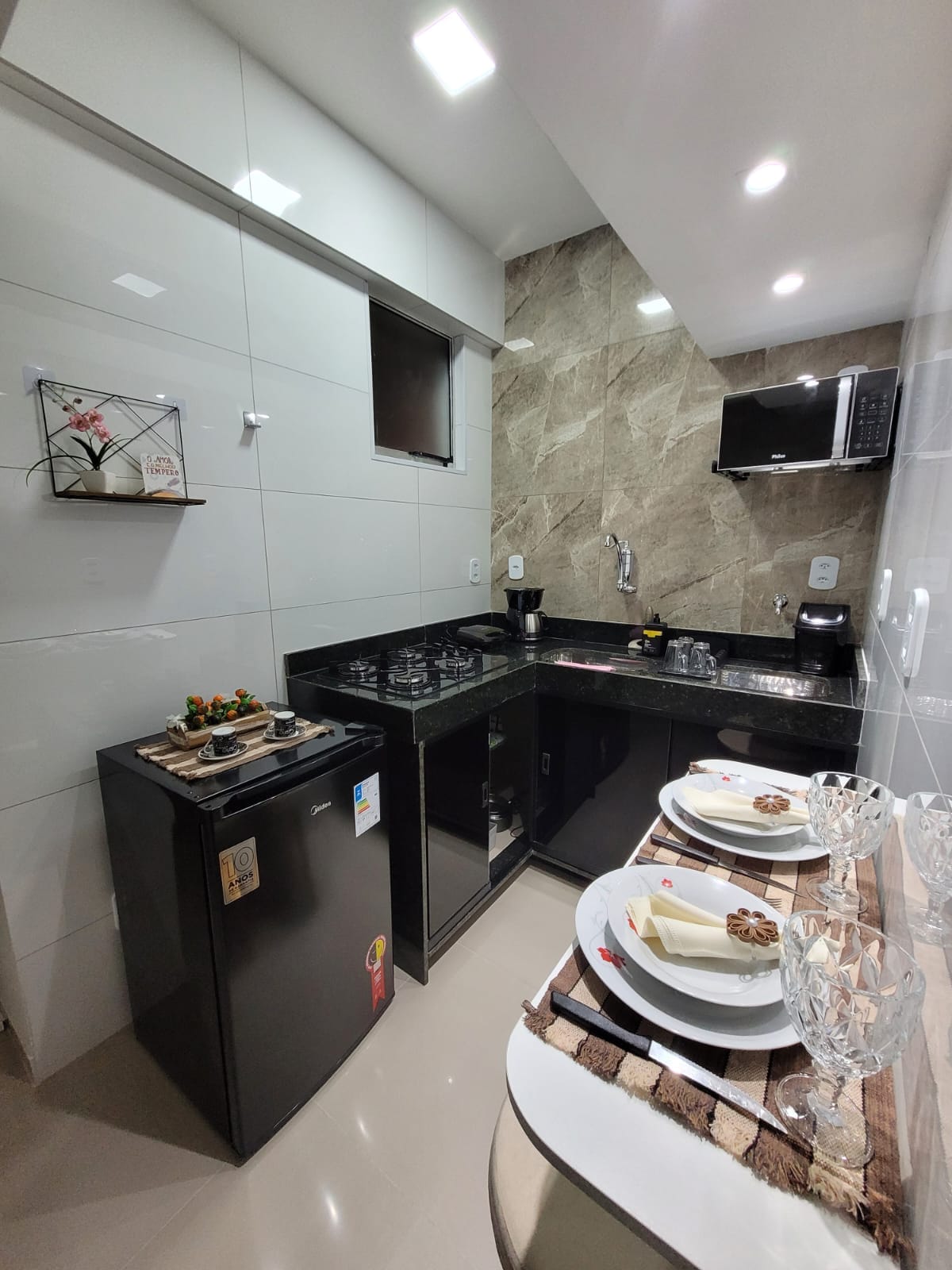 The kitchen features modern black countertops and cabinets, equipped with a refrigerator, stovetop, microwave, and sink. A dining area is set with neatly arranged tableware, including plates, glasses, and decorative items. Soft lighting enhances the contemporary design.