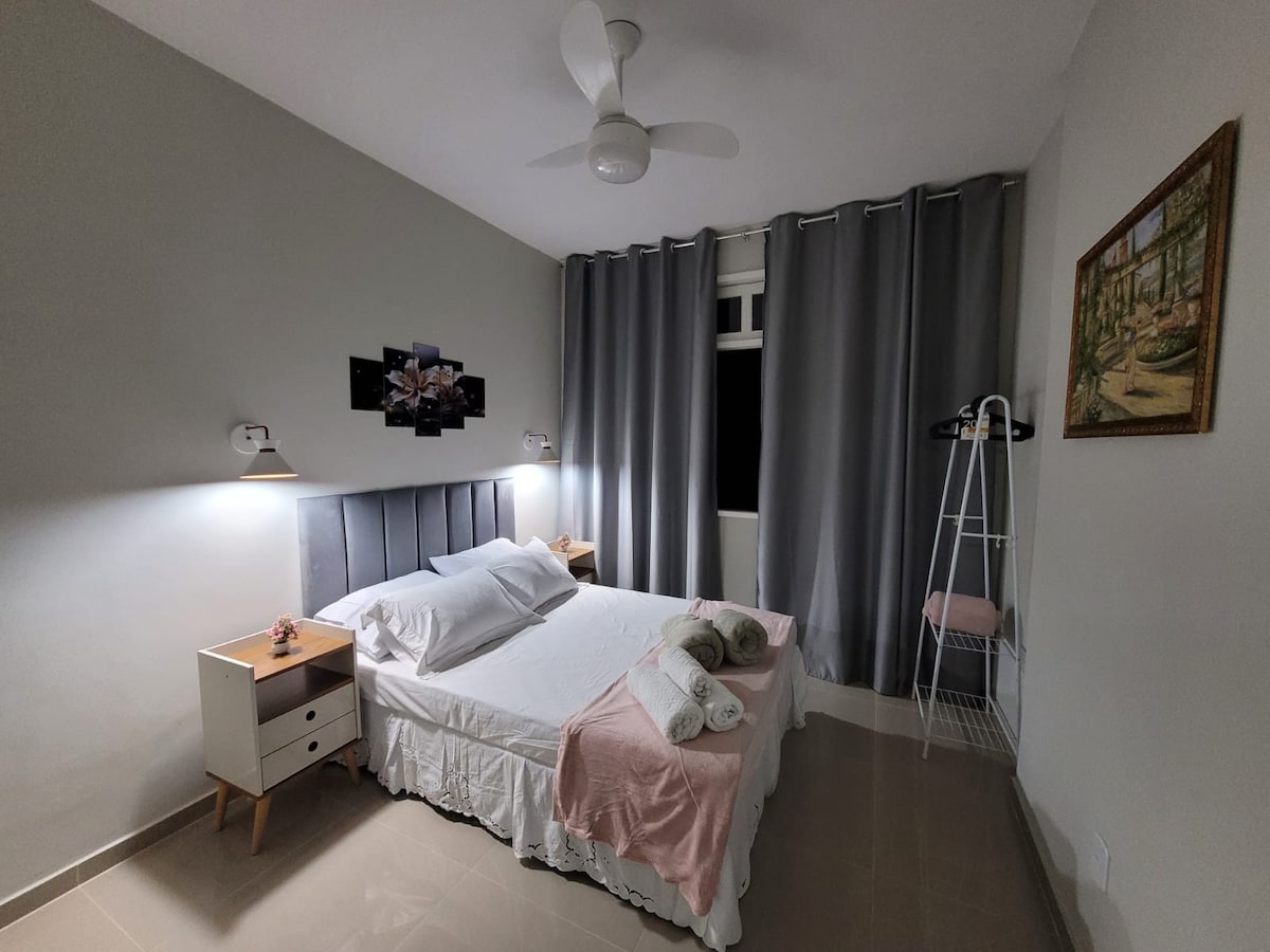 A welcoming bedroom features a spacious bed with light bedding and decorative pillows. Two bedside lamps provide soft lighting beside the bed. A ceiling fan enhances air circulation. Dark curtains frame the window, while a small shelf is positioned in the corner for added convenience.