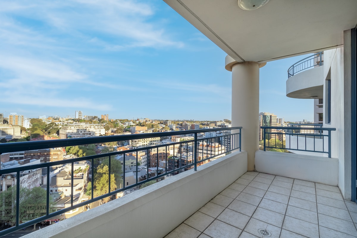 A spacious private balcony offers sweeping views of the surrounding cityscape. The tiled floor reflects natural light, while the railing ensures safety. The interior can be accessed through sliding glass doors, providing a seamless connection between indoor and outdoor spaces.