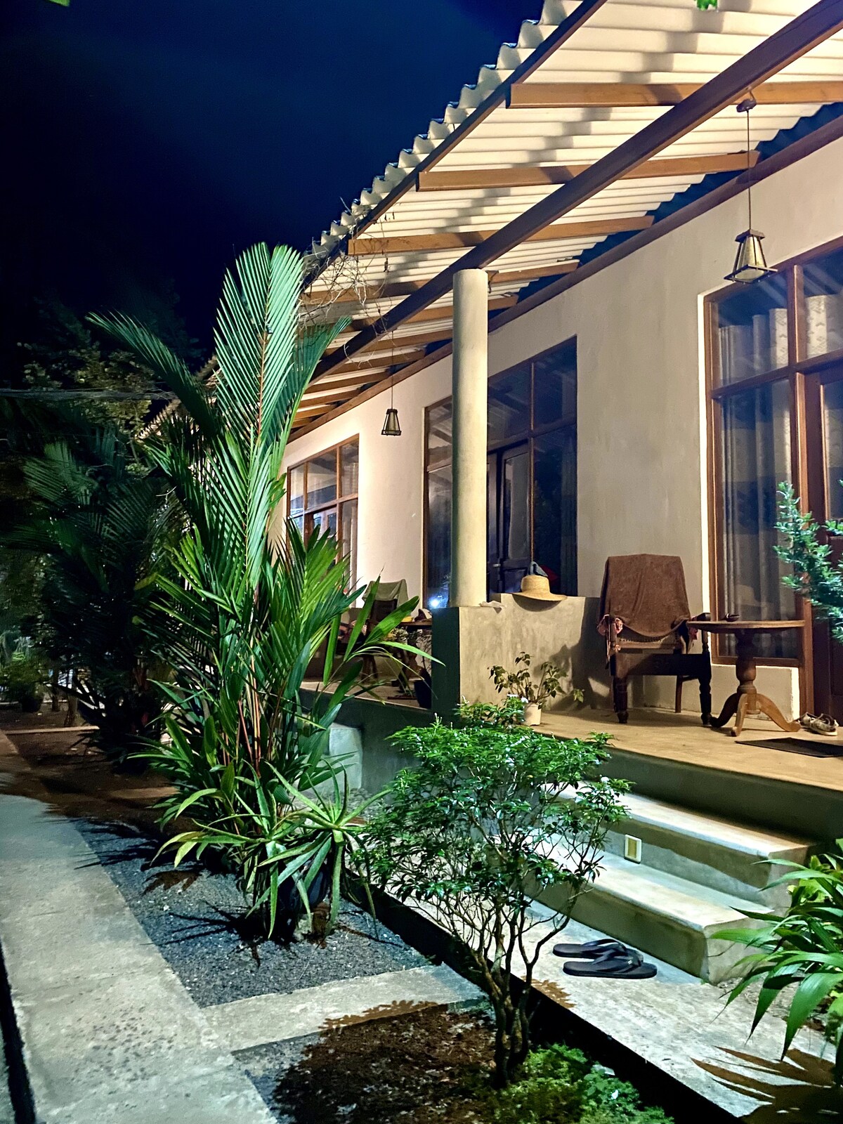 The exterior of a charming retreat is illuminated at night, showcasing a porch with a wooden chair and a decorative item. Lush greenery, including palms and shrubs, surrounds the entrance, enhancing the serene atmosphere of the secluded setting.