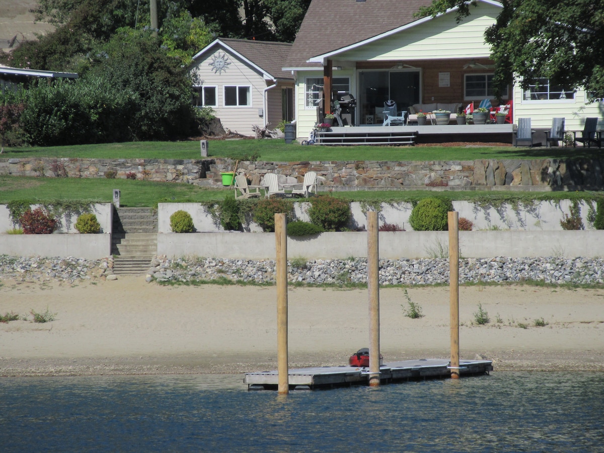 A sandy beach forms the foreground, leading to a low dock supported by wooden posts. In the background, a two-story house with a covered porch is visible, surrounded by greenery and landscaping. Stairs connect the beach area to the yard, enhancing accessibility.