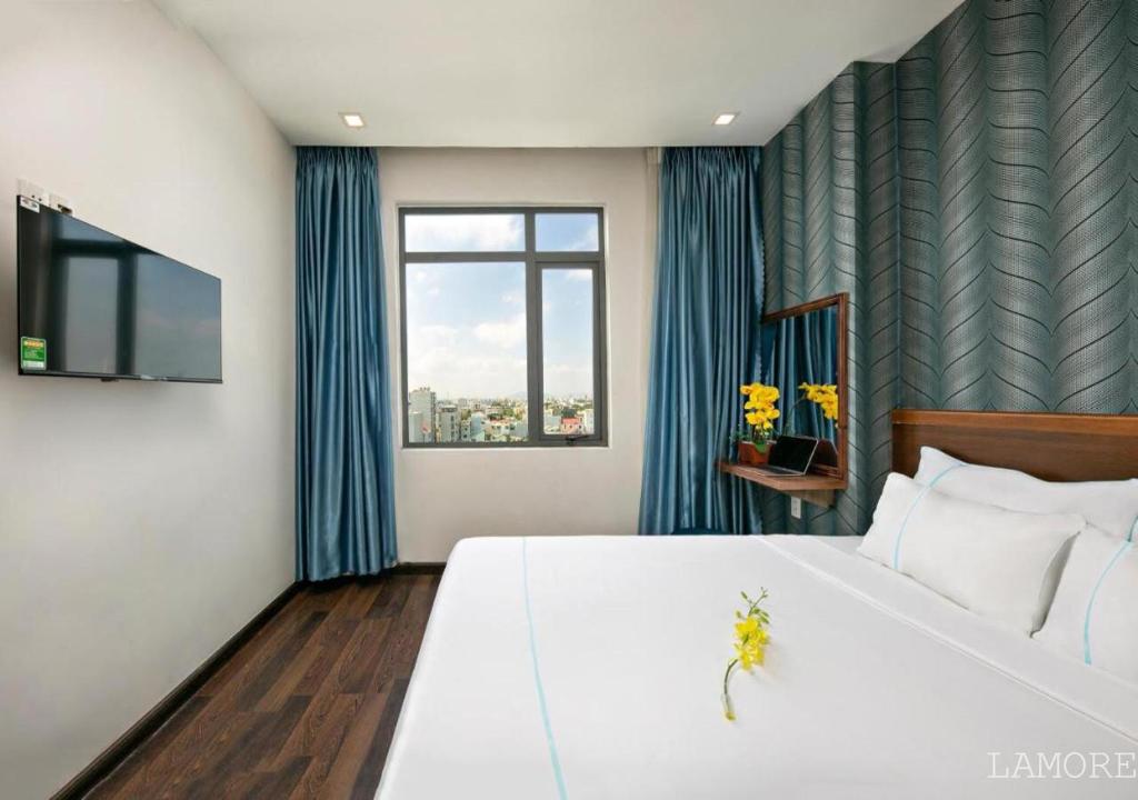 A bright room features large windows offering city views, with natural light illuminating the space. A comfortable bed is dressed in fresh linen, complemented by decorative elements such as a small flower. A flat-screen TV is mounted on the wall, adding convenience.