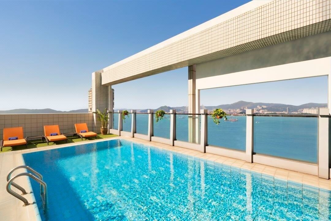 A rooftop pool is framed by modern tiling and glass panels, offering panoramic views of the water and distant mountains. Vibrant orange lounge chairs are positioned beside the pool, creating a spacious area for relaxation under a clear blue sky.