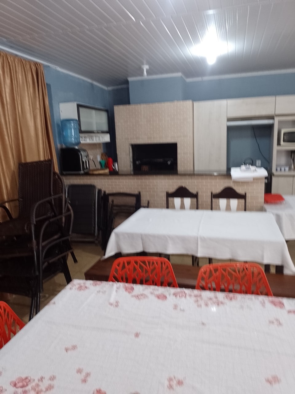 A dining area is present with several tables set with white tablecloths, accompanied by colorful chairs. A well-equipped kitchen can be seen in the background, featuring modern appliances and a counter, while a few stacked chairs indicate the space's versatility.