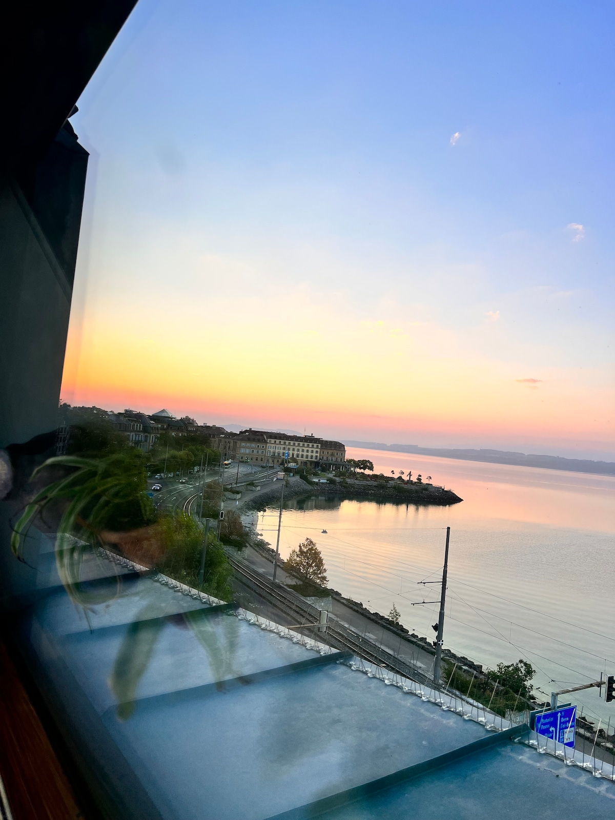 A scenic view is captured through a large window, showcasing a calm lake at sunset. Soft colors in the sky blend with the water, while a curving road and buildings line the lakeshore, contributing to a tranquil ambiance.