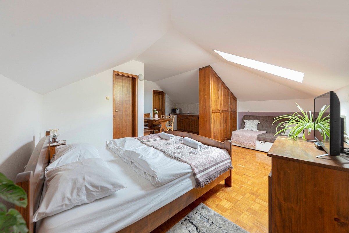 An airy attic room features a spacious bed with white linens and a patterned blanket. Natural light filters through a skylight, illuminating the wooden furniture, including a dresser and a seating area with a sofa. A small kitchenette can be seen in the background.