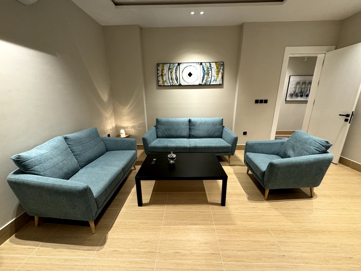 A cozy living area features two light blue sofas, one armchair, and a black coffee table at the center. Soft lighting highlights a piece of wall art above the seating, while a side table with a decorative item adds to the inviting atmosphere.